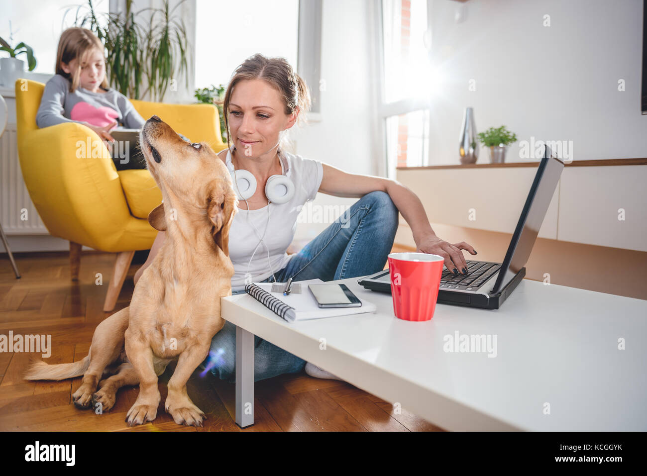 Femme assise à la table à café sur le sol et caresser chien à la maison tandis que sa fille assise sur le fauteuil et l'utilisation de tablet Banque D'Images Femme assise à la table à café sur le sol et caresser chien à la maison tandis que sa fille assise sur le fauteuil et l'utilisation de tablet Banque D'Images