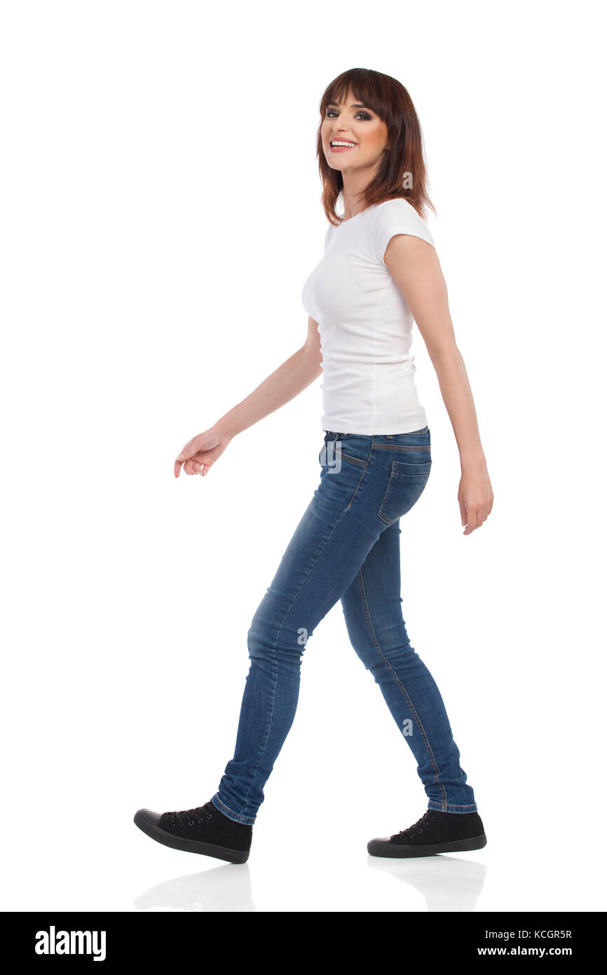 Jeune femme en jeans et t-shirt blanc est la marche, looking at camera and smiling. full length portrait isolé sur blanc. Banque D'Images