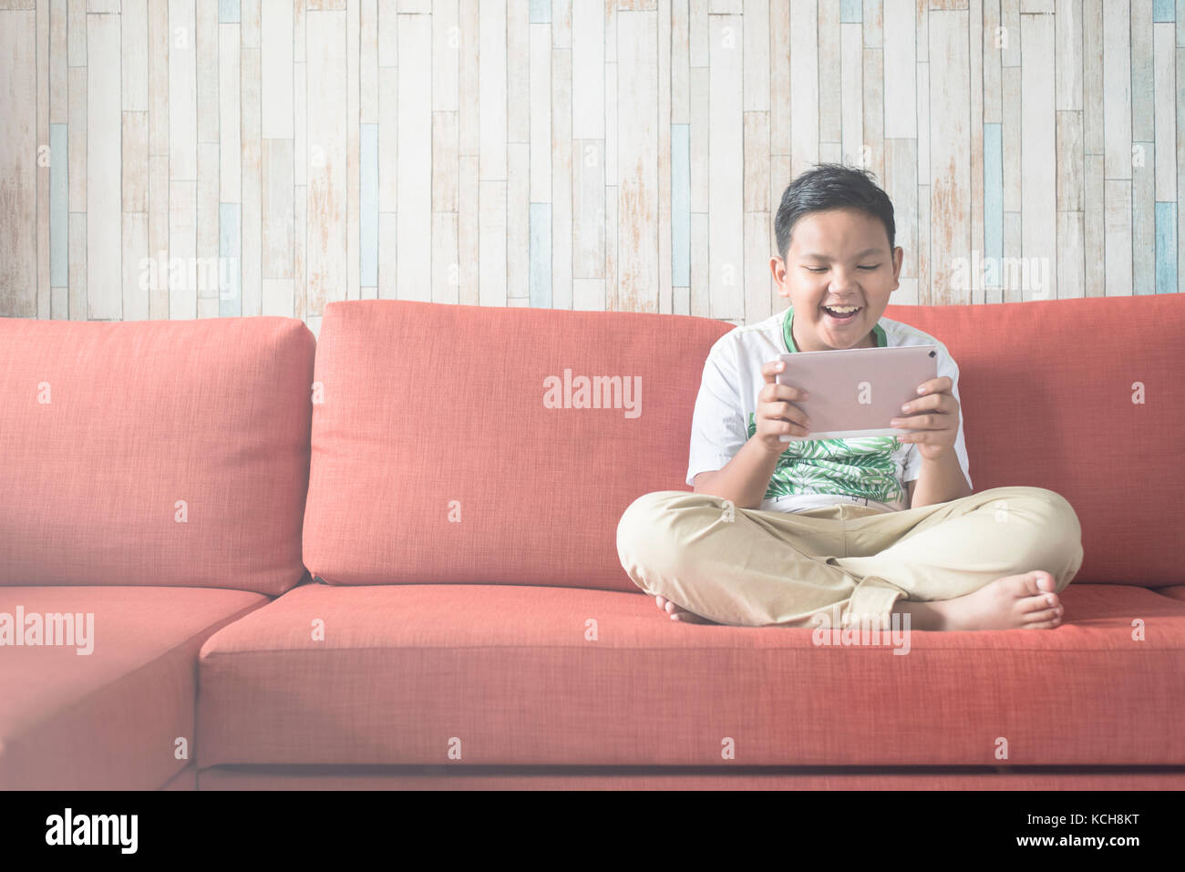 Young Asian boy using digital tablet sur un canapé à la maison assis en tailleur. boy walking. technologie / concept d'apprentissage sur ordinateur il. Banque D'Images