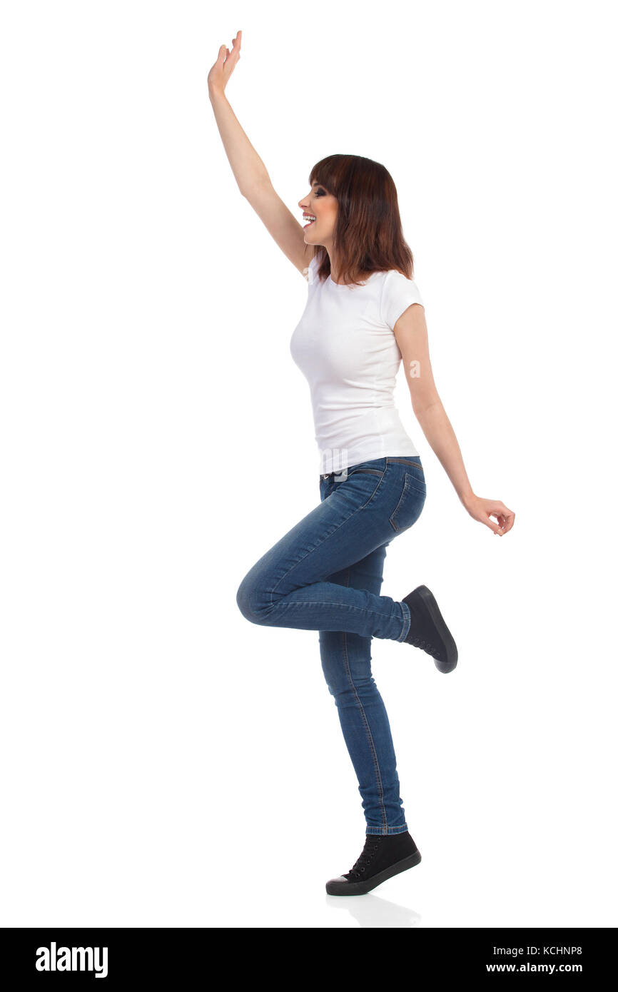Jeune femme en jeans et t-shirt blanc est debout sur une jambe, l'augmentation des bras, forme, à l'écart et crier. full length portrait isolé o Banque D'Images