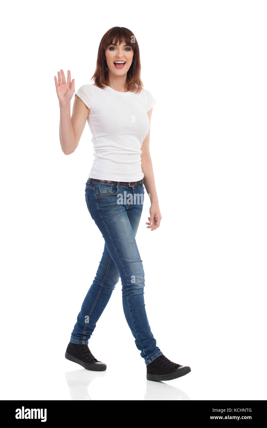 Jeune femme en jeans et t-shirt blanc est la marche, looking at camera, agitant la main et parler. full length portrait isolé sur blanc. Banque D'Images