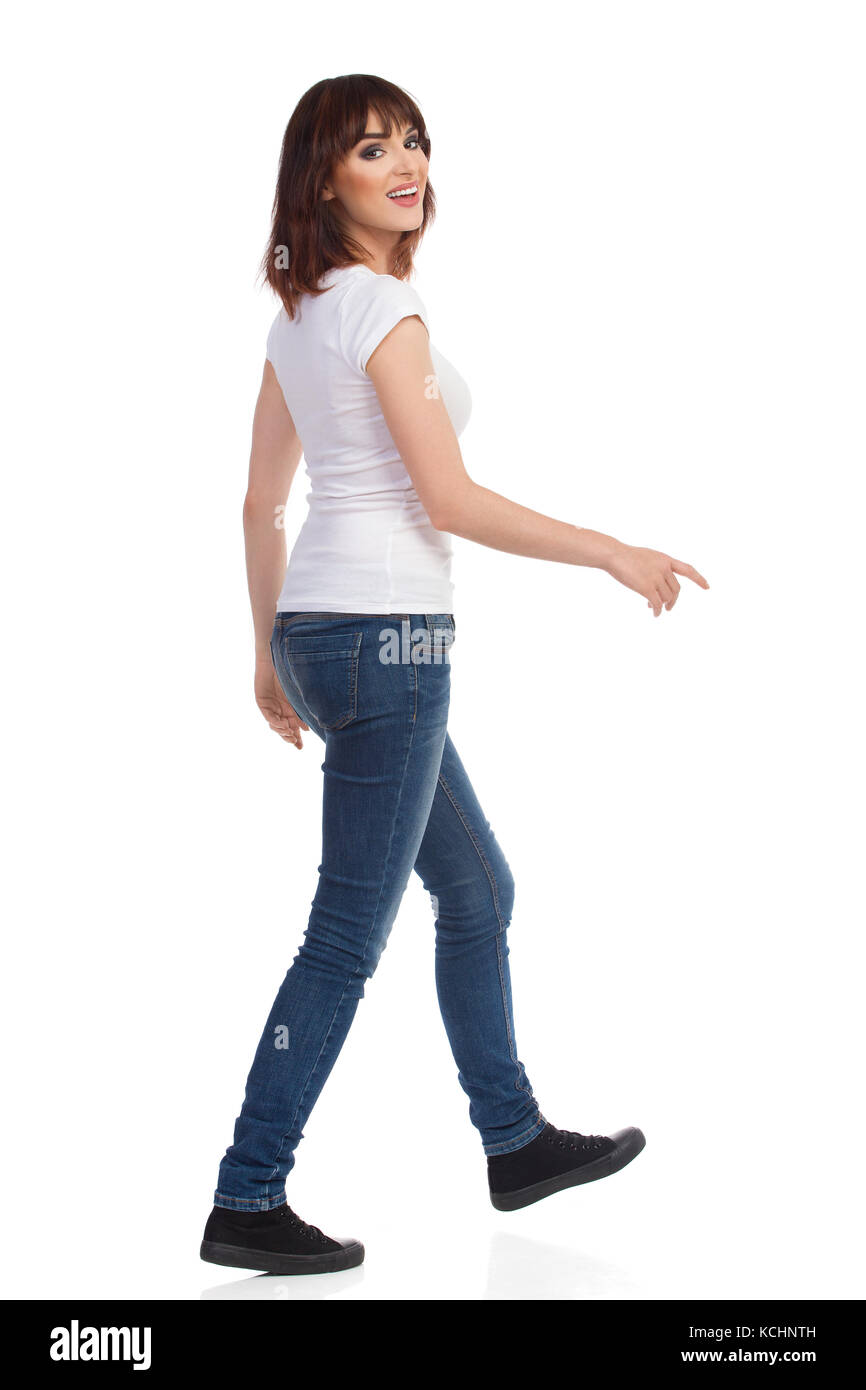 Jeune femme en jeans et t-shirt blanc est la marche, à la caméra au-dessus de l'épaule et parler. full length portrait isolé sur blanc. Banque D'Images
