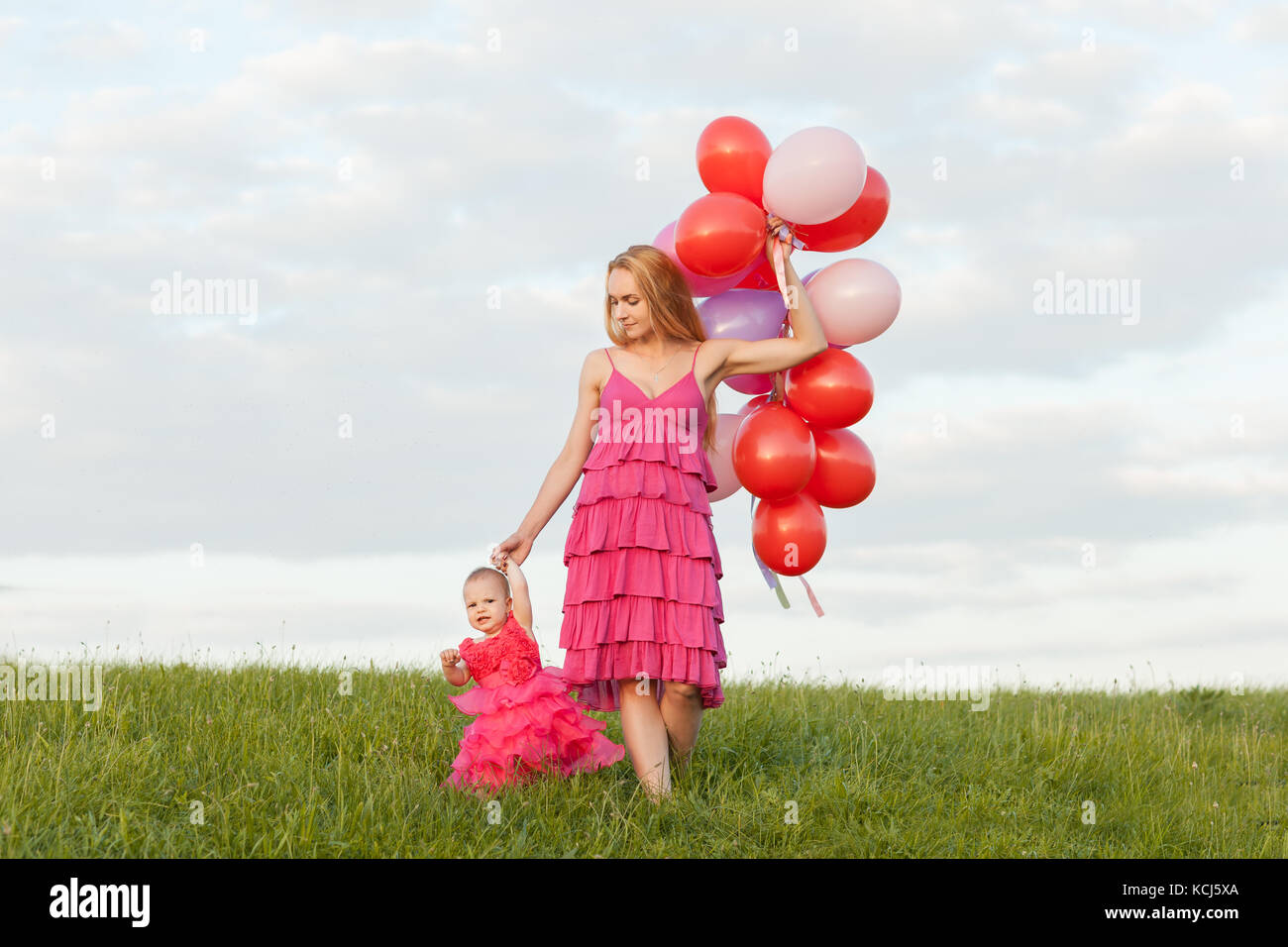 Mère et fille dans de belles robes sont à pied le long de l'herbe verte. maman est titulaire d'un tas de ballons dans ses mains Banque D'Images