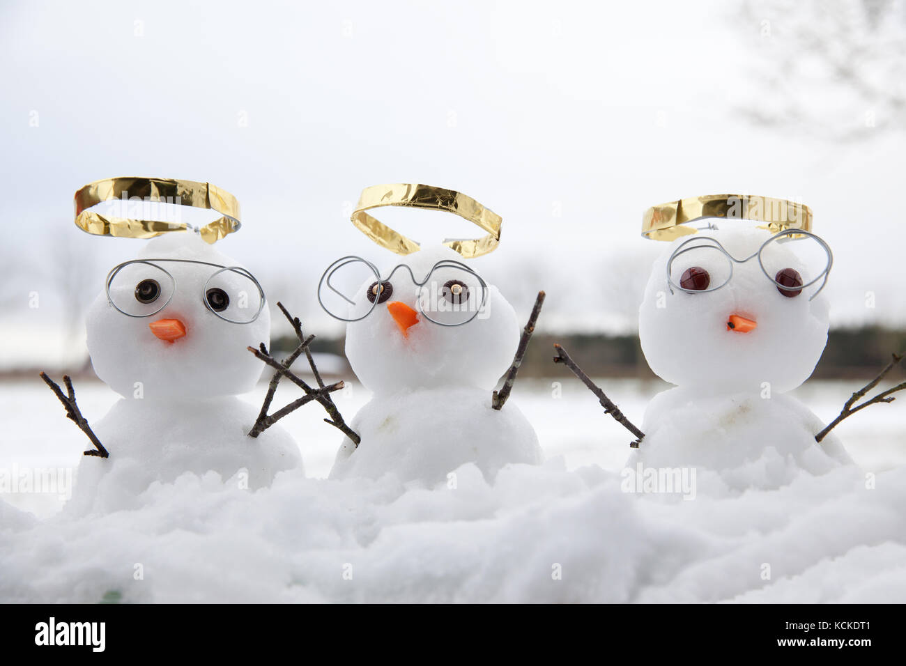 Trois angles bonhomme mignon avec golden halos et lunettes de lecture et leurs rameaux en bois les bras en l'air. la neige tomber sur le sol en hiver Banque D'Images