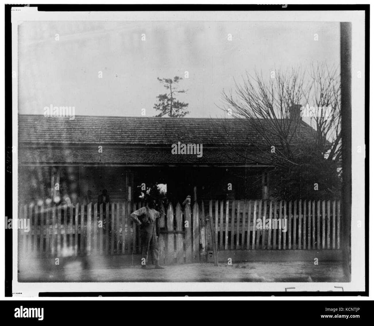 Cette photographie montre un homme afro-américain debout près d'une grande clôture de piquet devant une maison, capturant un moment dans le temps qui met en valeur la vie rurale ou suburbaine. Banque D'Images