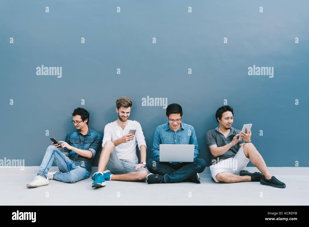 Groupe multiethnique de quatre hommes à l'aide de l'ordinateur portable, smartphone, tablette numérique avec copie espace sur le mur bleu. style de vie avec concept gadgets Banque D'Images