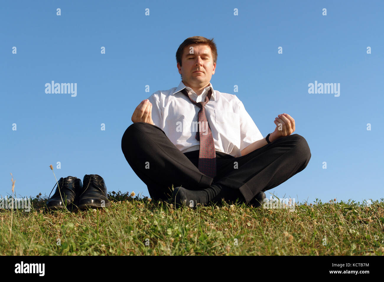 Businessman wearing chemise blanche et cravate en méditant sur l'herbe avec ses chaussures plus de ciel bleu clair Banque D'Images