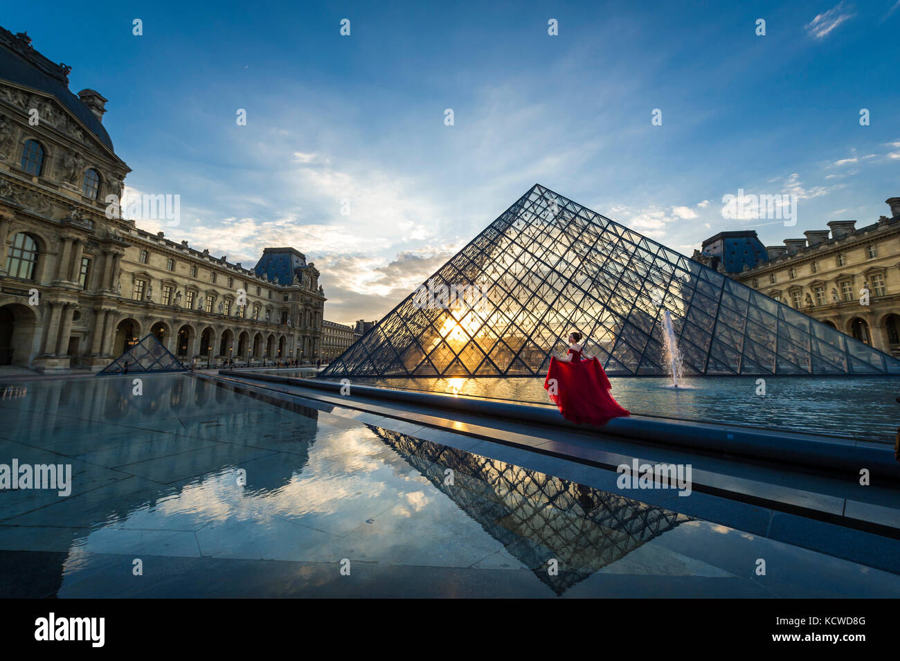 Modèle dans une robe rouge à la Pyramide du Louvre au coucher du soleil Banque D'Images
