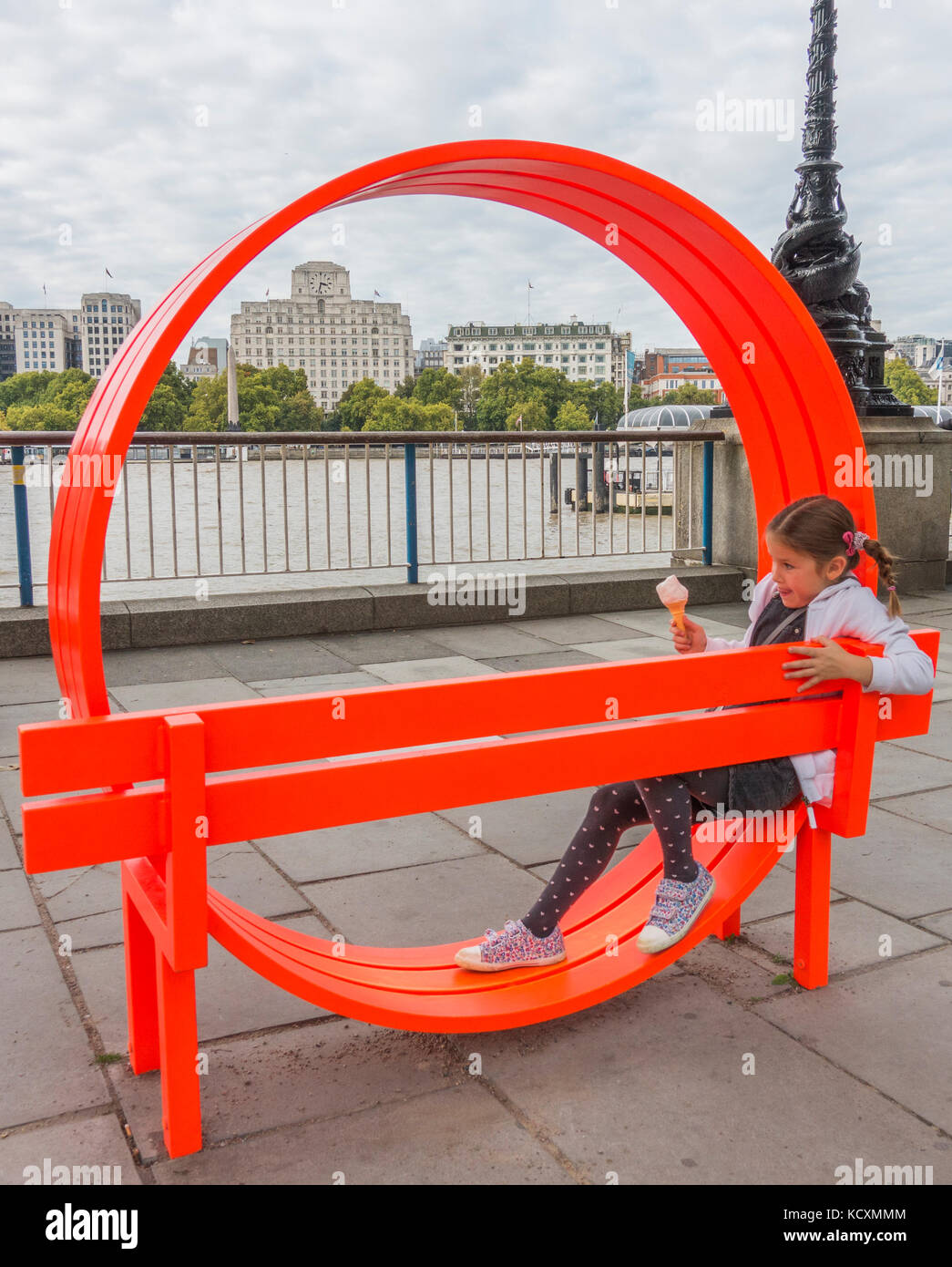 Une jeune fille tenant un cornet de crème glacée tout en jouant sur un banc orange distinctive sur rive sud, donnant sur la Tamise, Londres, Angleterre, Royaume-Uni. Banque D'Images