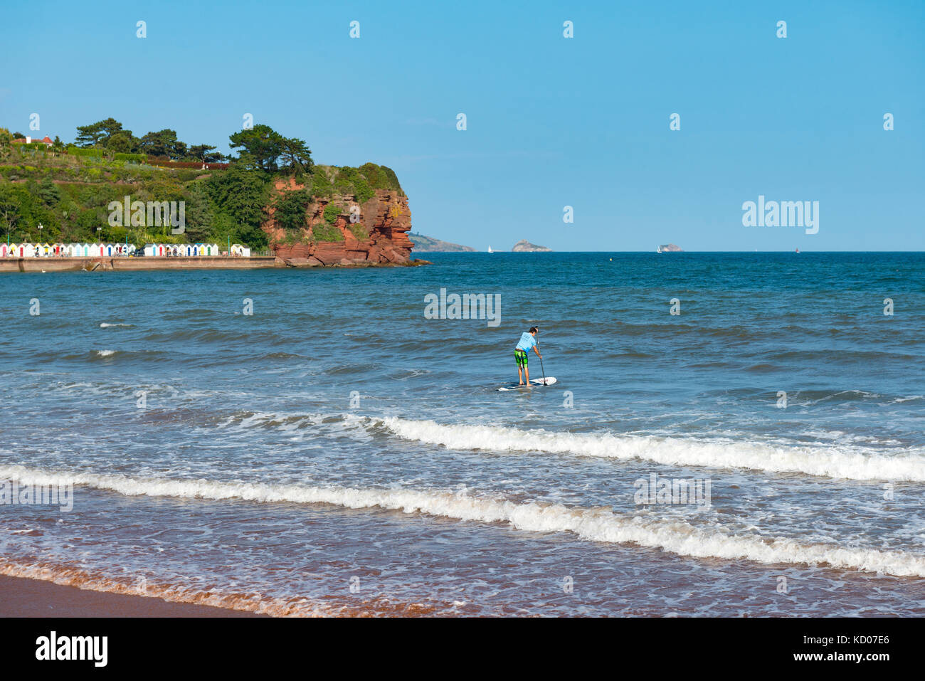 Paddle-boarder de partir à la mer à plage de goodrington sands, Paignton, Devon. uk. cabanes de plage et falaises rouges à tête roundham avec bateau dans la distance Banque D'Images