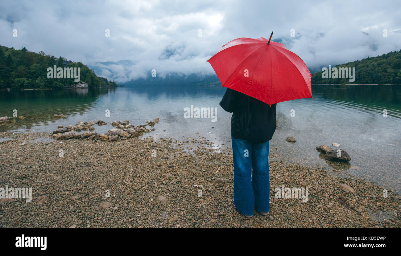 Femme avec parapluie rouge prévoit de la pluie en face d'un lac. triste et solitaire à la personne de sexe féminin dans la distance. Banque D'Images