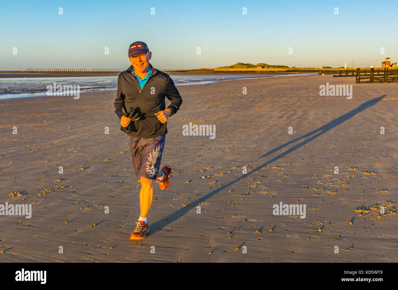 Man smiling at the camera tout en ayant un jogging du matin sur une plage de sable fin, illuminé par le soleil du matin, au Royaume-Uni. Banque D'Images