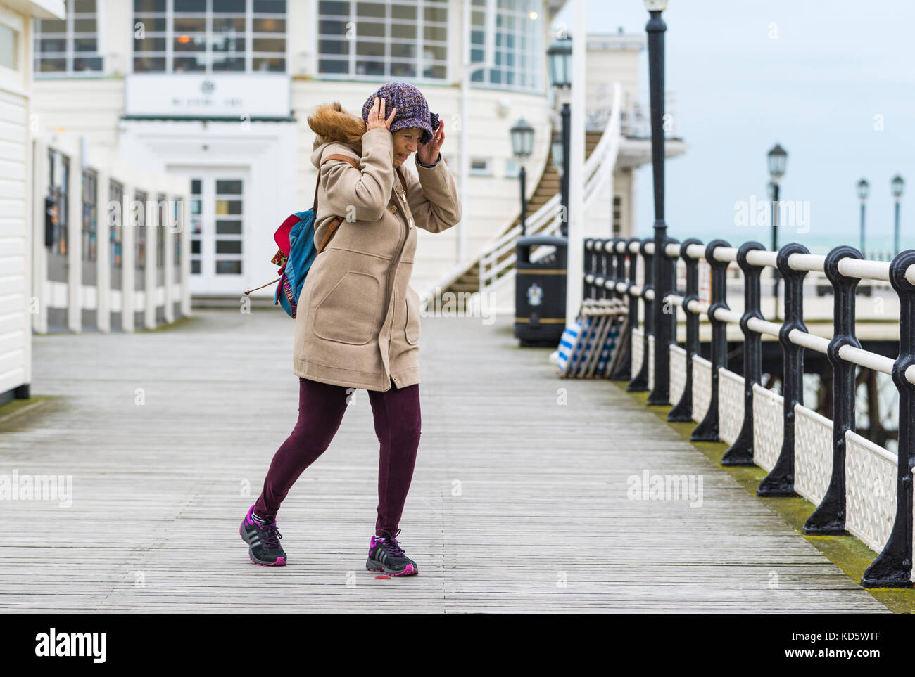 Femme de la difficulté à marcher dans le vent tout en tenant son chapeau sur la jetée de Worthing à Worthing, West Sussex, Angleterre, Royaume-Uni. Banque D'Images