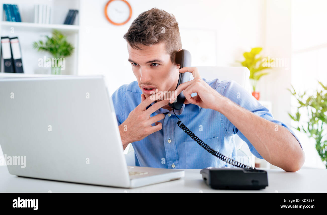Beau jeune homme pensif avec phone and looking at laptop dans le bureau. Banque D'Images