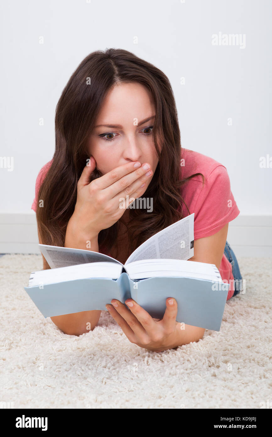 Choqué young woman lying on carpet reading book Banque D'Images