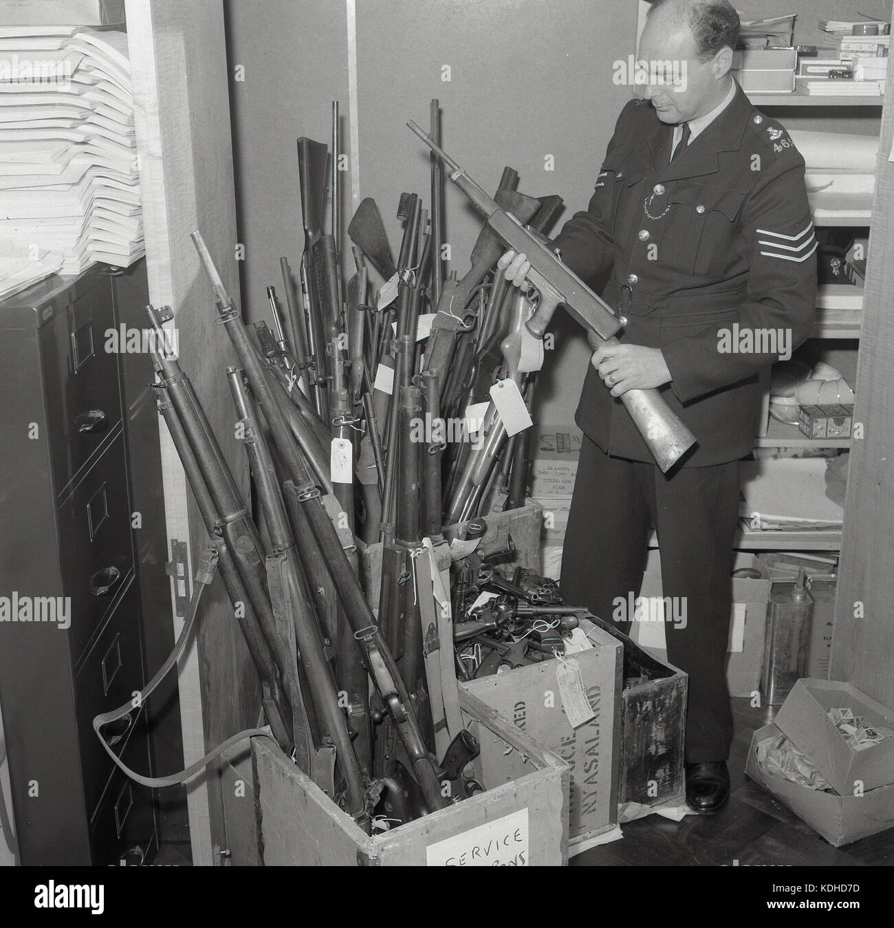 Années 1960, historiques, sergent de la police britannique mâle tenant un fusil, sélectionnés à partir d'une cachette de fusils, pistolets et autres armes similaires debout dans des boîtes en bois, Buckinghamshire, Angleterre, Royaume-Uni. Les Anglais forcedepolice fut à cette époque une force non armée et encore aujourd'hui. Banque D'Images