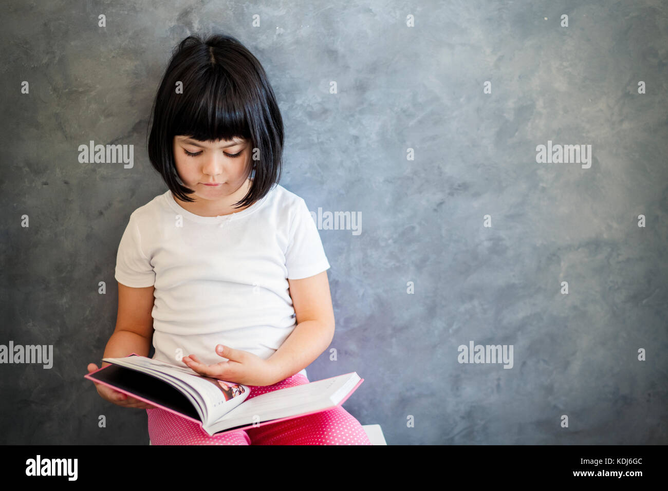 Portrait of cute little girl cheveux noir livre de lecture par le mur Banque D'Images