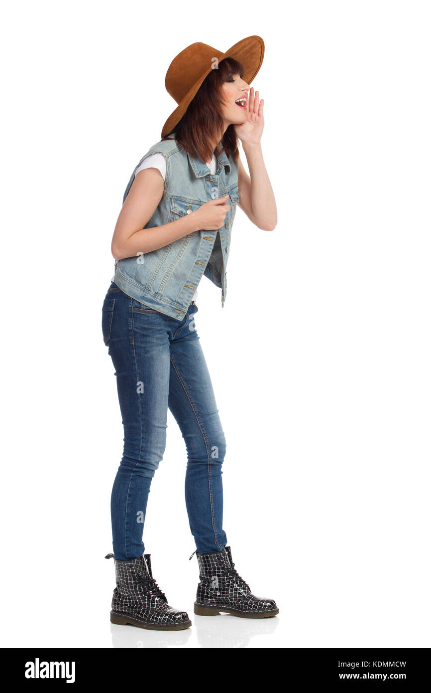 Jeune femme en jeans gilet, bottes noires et brown suede hat est debout, tenant la main sur le menton et crier. Vue de côté. toute la longueur isolée studio shot Banque D'Images