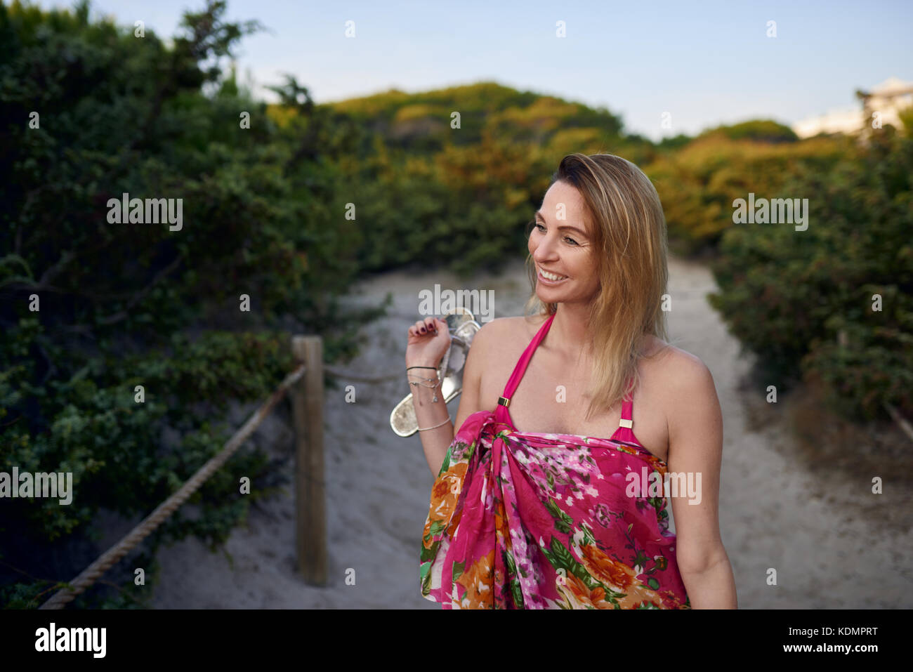 Jolie femme mince transportant ses chaussures aux pieds nus comme elle marche le long d'une plage de sable fin sourire joyeusement à chemin l'appareil photo en vacances Banque D'Images