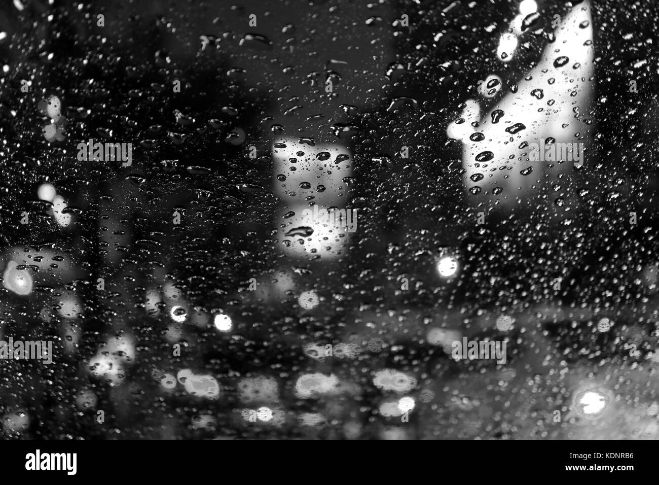 Photo monochrome des lumières de rue floue et les feux arrière vu à travers les gouttes de pluie sur le pare-brise de voiture dans la nuit Banque D'Images