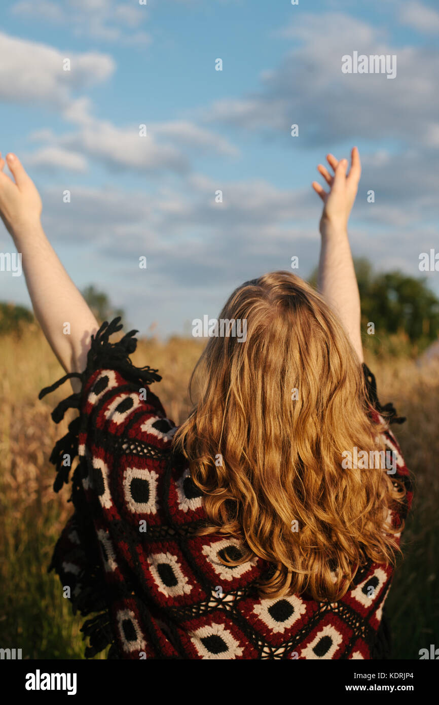 Le dos d'une jeune femme avec de longs cheveux blonds d'élever ses bras vers le ciel dans un champ, Norfolk, uk Banque D'Images