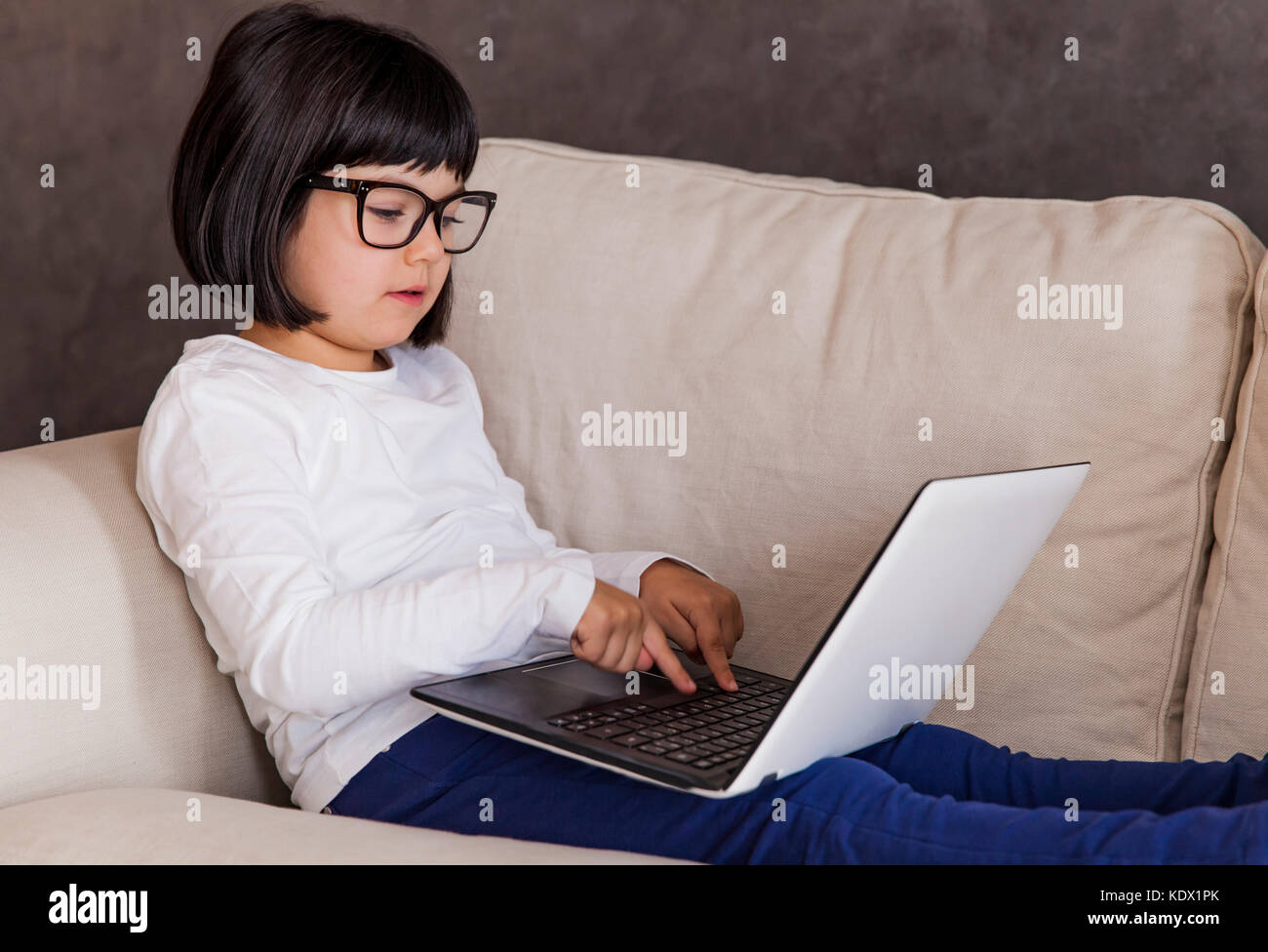 Petite fille aux cheveux noirs à l'aide de son ordinateur portable tout en lunettes assis sur le canapé à la maison Banque D'Images