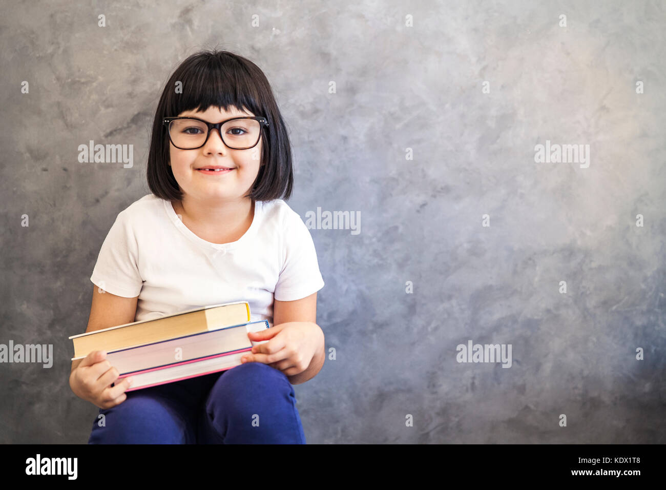 Portrait of smiling black hair petite fille avec des lunettes holding books par le mur Banque D'Images