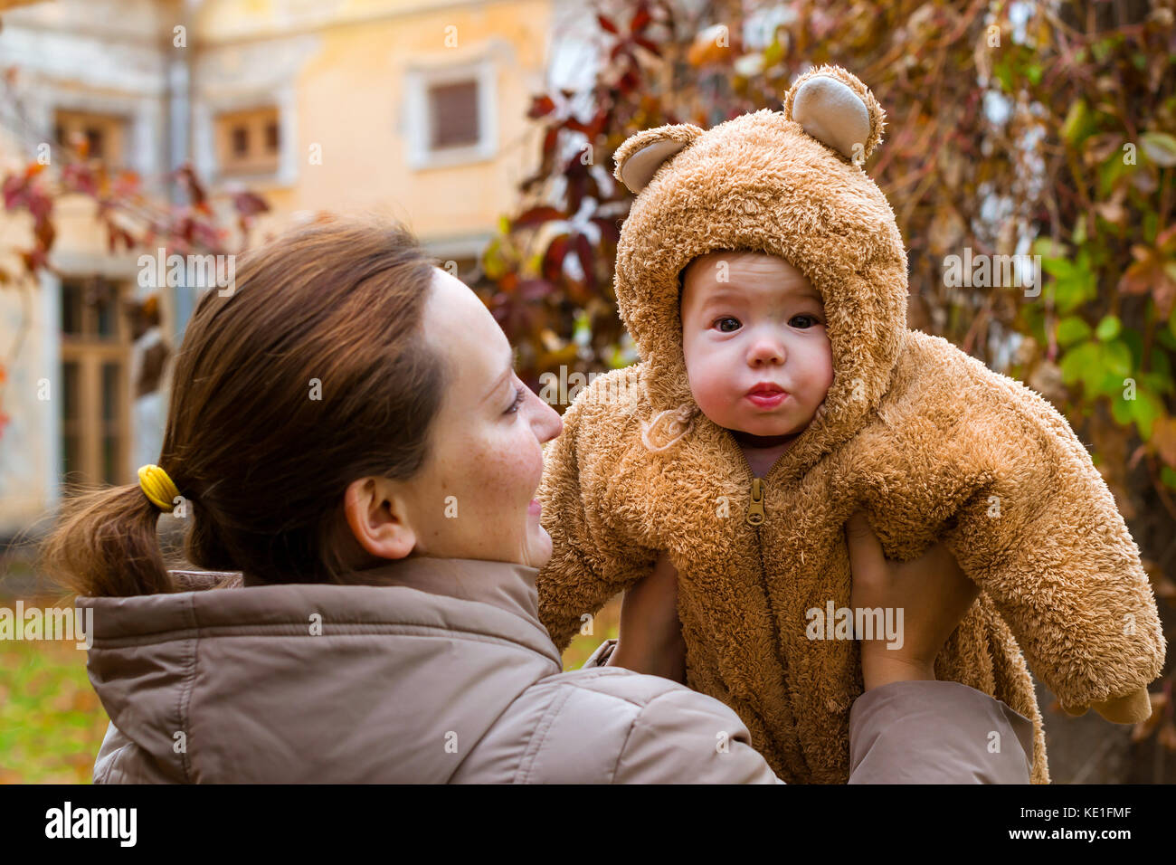 Femme avec bébé dans les bras, posant sur fond d'automne parc. enfant vêtu de teddy-bear stylisé chaud costume, s'amusant sur les bras de la mère de famille. w Banque D'Images