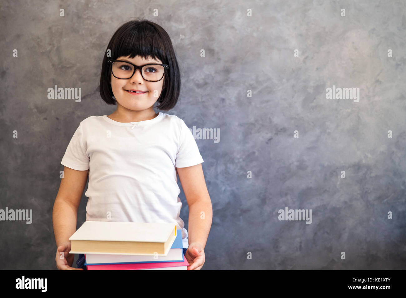 Portrait of smiling black hair petite fille avec des lunettes holding books par le mur Banque D'Images
