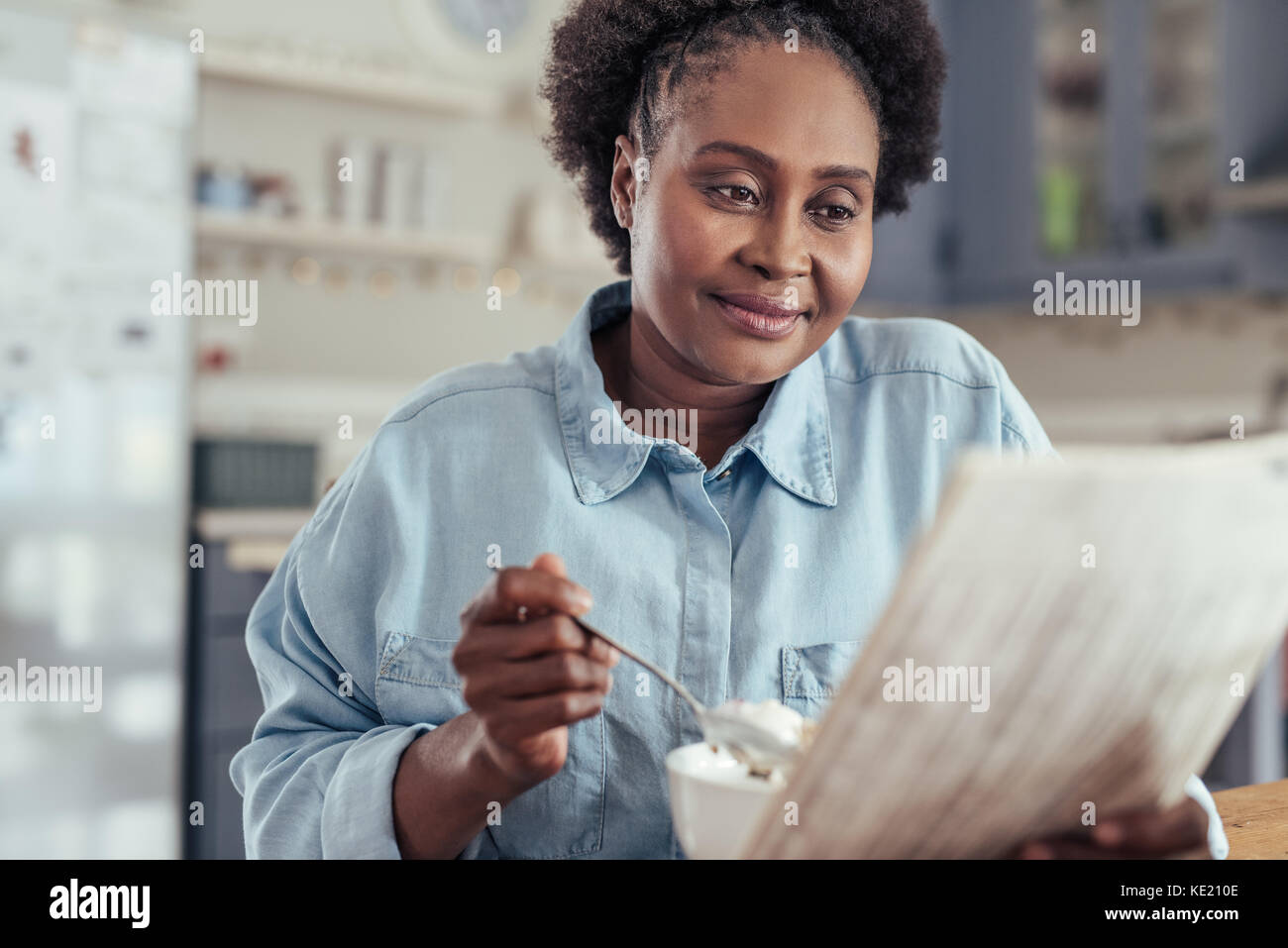 Jeune femme africaine assis à sa table de cuisine le matin, lire le journal et manger un petit déjeuner sain Banque D'Images