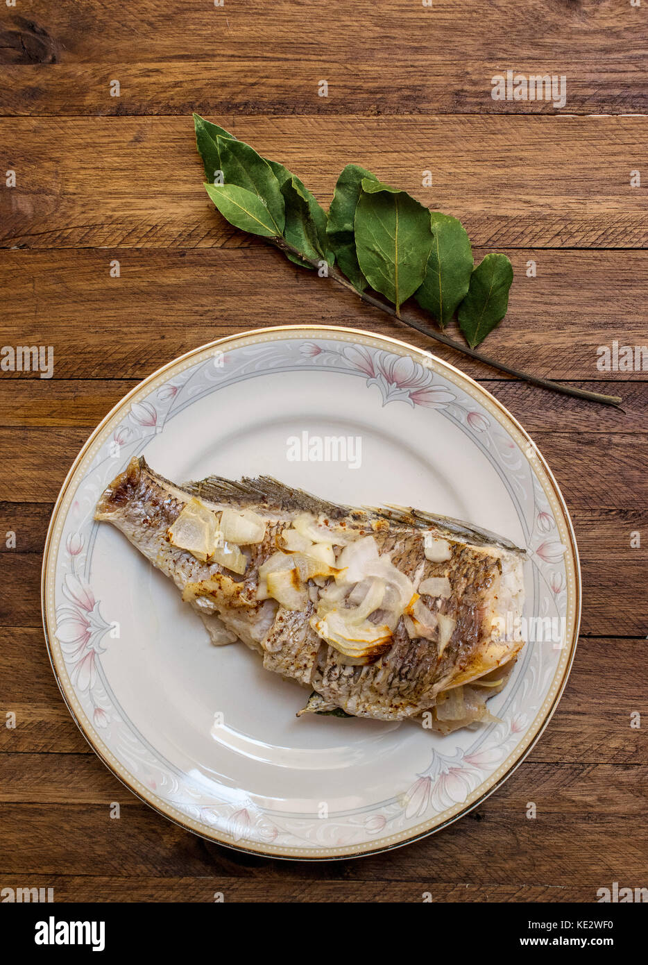 Le poisson entier, cuit au four avec ail et oignon tranché sur le dessus, sur une plaque décorative en porcelaine blanche, sur un fond de bois, avec une feuille de laurier bran Banque D'Images