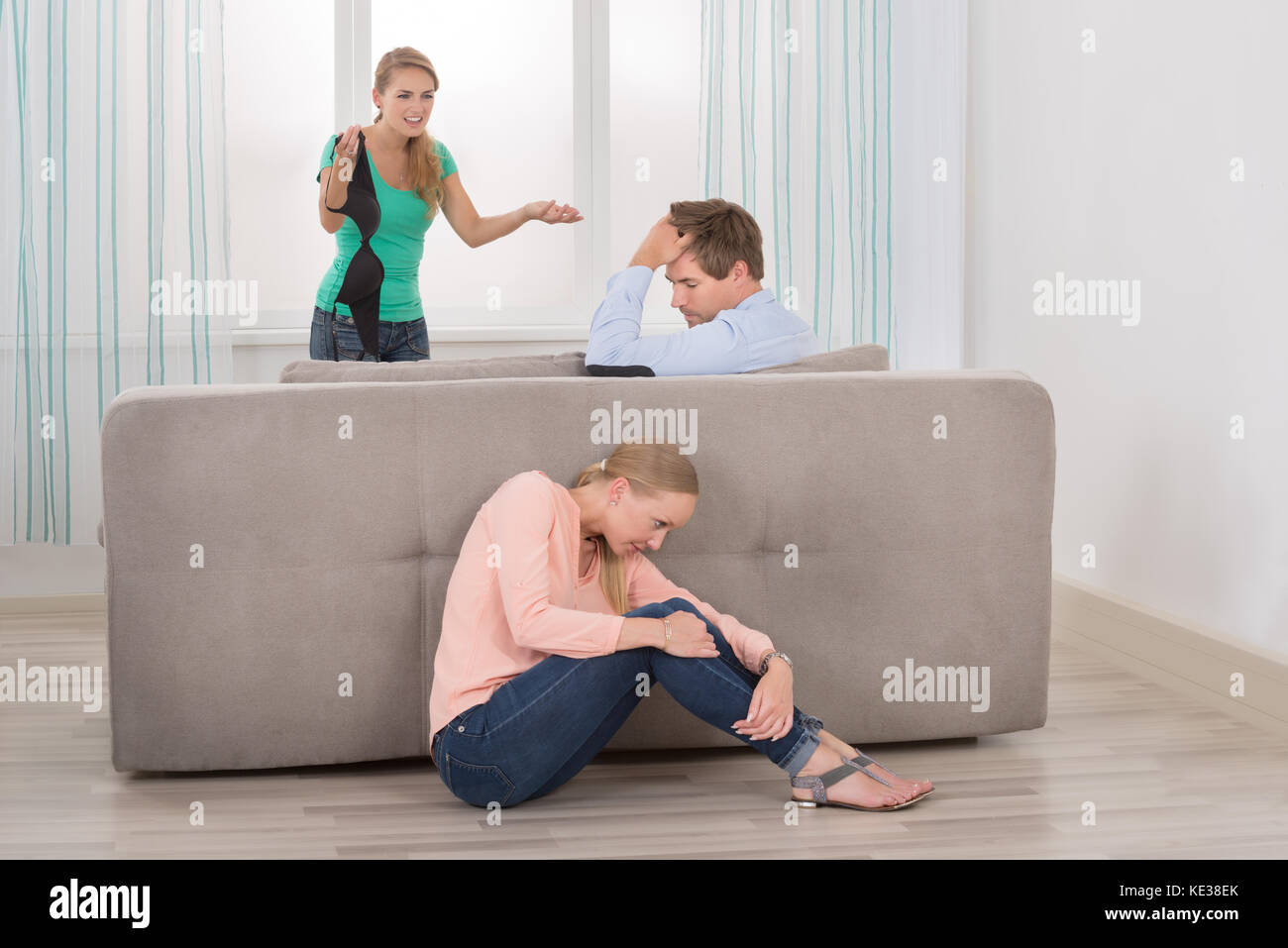 Wife Holding Bra se disputer avec son mari alors que sa petite amie se cacher derrière un canapé Banque D'Images