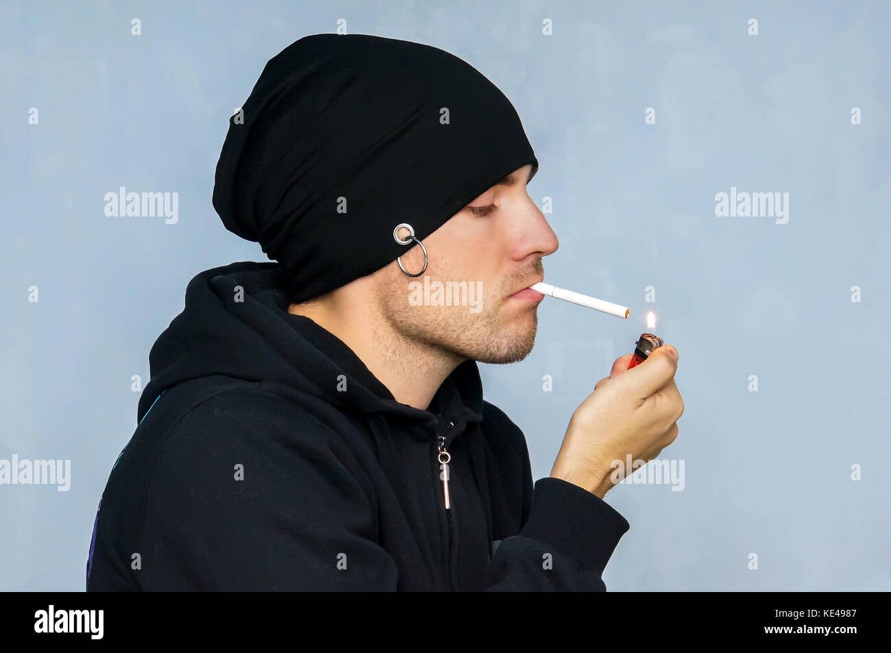 Jeune fumeur dans un chapeau noir il allume une cigarette à partir de l'allume-cigare sur un fond bleu. punk fume. vêtements sous-culturelle d'un toxicomane. Banque D'Images