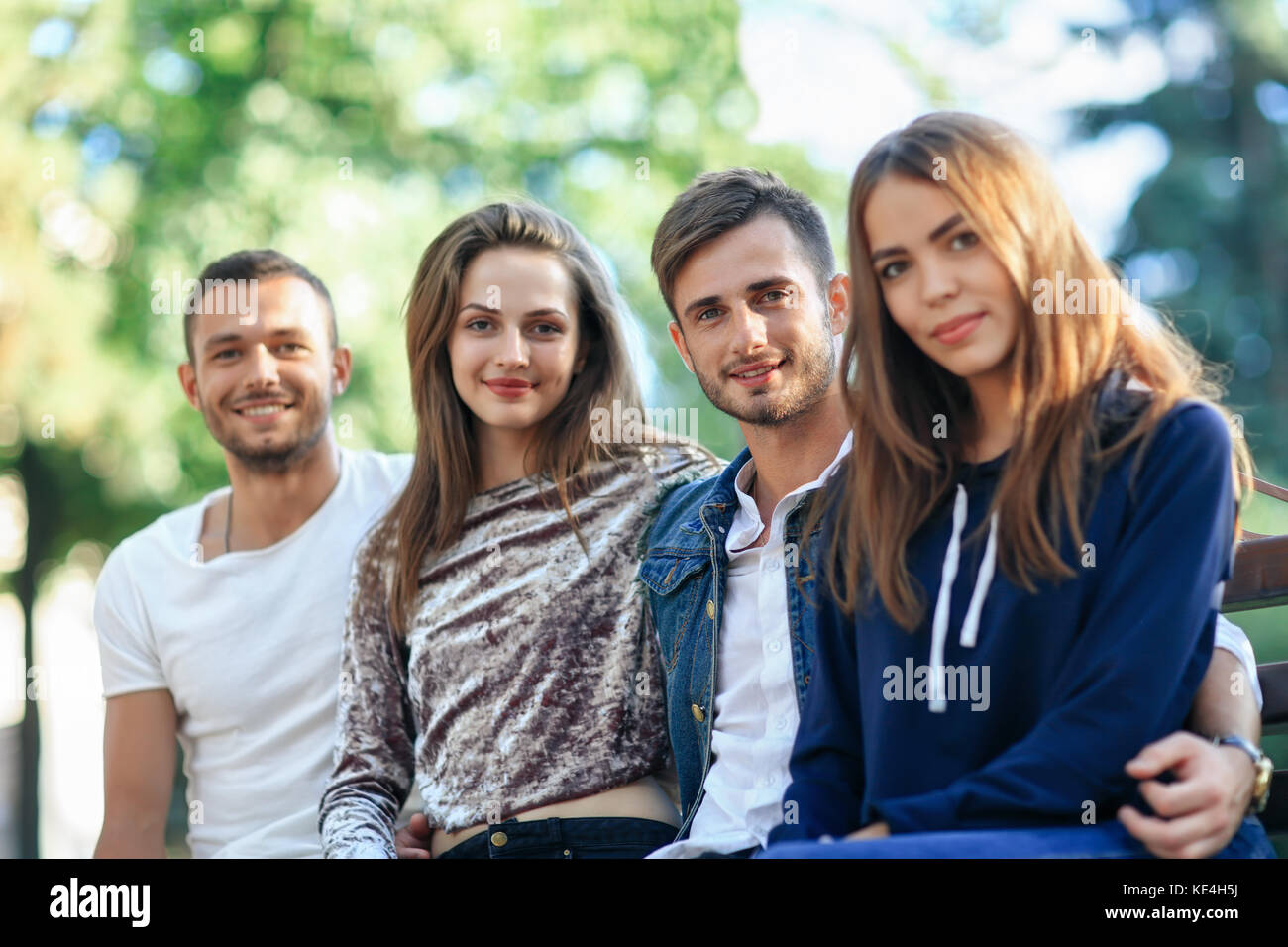 Quatre femmes et hommes assis sur un banc à l'embrasser. heureux jeune élégant friends posing on bench outdoors, smiling at camera et en bonne santé. Banque D'Images