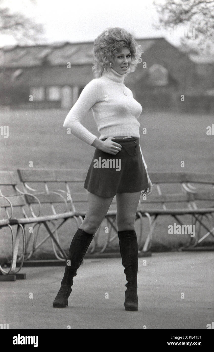 Années 1960, historiques, femme blonde bien roulée avec de grands cheveux ondulés posant dehors dans une mini jupe noire et des bottes aux genoux. Banque D'Images