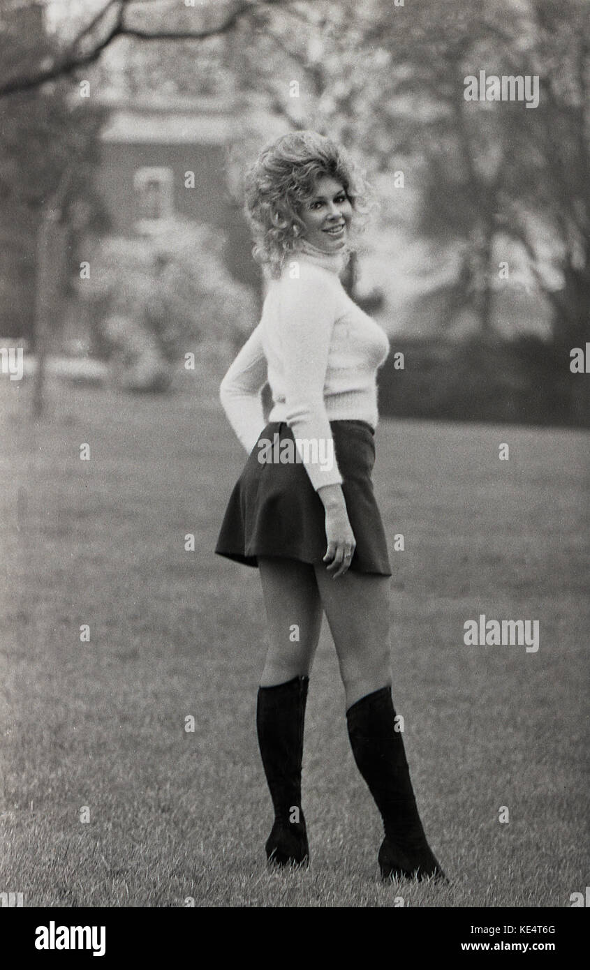 Années 1960, historiques, femme blonde bien roulée avec de grands cheveux ondulés posant dehors dans une mini jupe noire et des bottes aux genoux. Banque D'Images