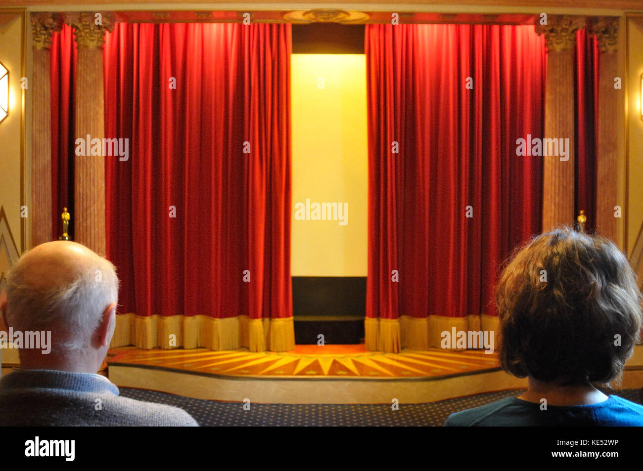 Des rideaux rouges de velours légèrement ouverte montre homme et femme regardant dans salle de cinéma construit en 1920, début des années 1930 style. Bien éclairée. Banque D'Images
