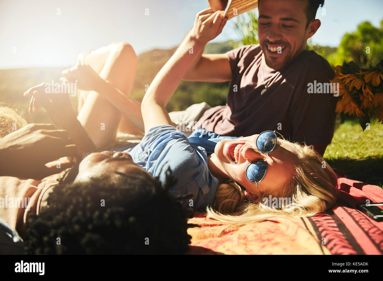De jeunes amis ludiques rient, se détendent sur une couverture de pique-nique dans un parc ensoleillé d'été Banque D'Images