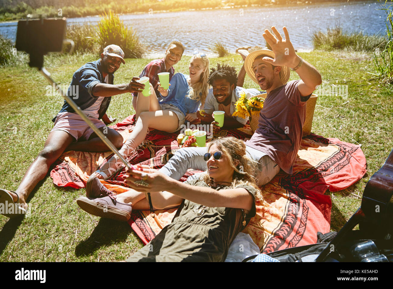 De jeunes amis joueurs prennent le selfie avec un bâton de selfie, en appréciant un pique-nique au bord de la rivière ensoleillée en été Banque D'Images