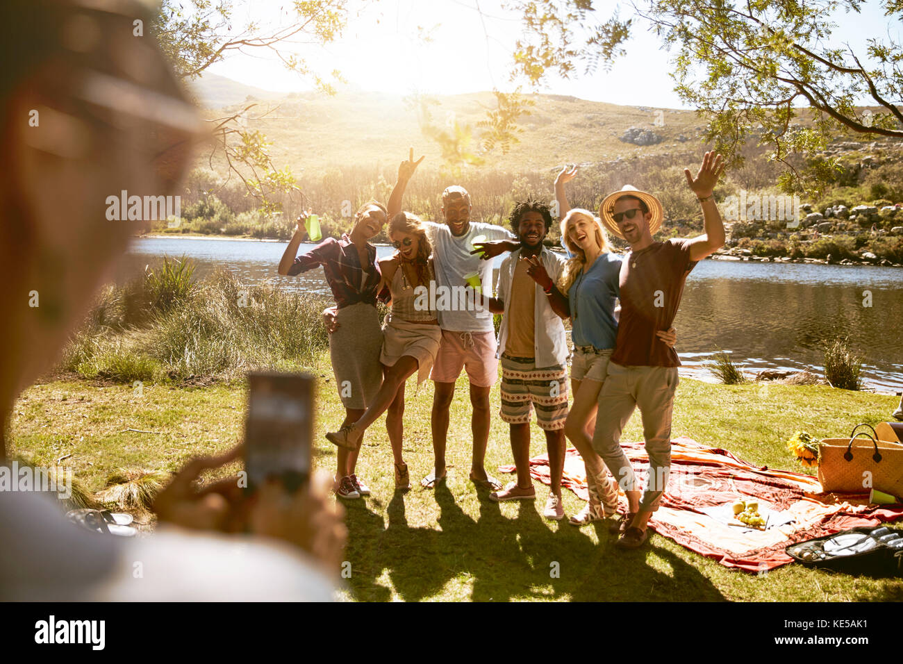 Jeune femme avec appareil photo téléphone photographier des amis au soleil de l'été pique-nique au bord de la rivière Banque D'Images
