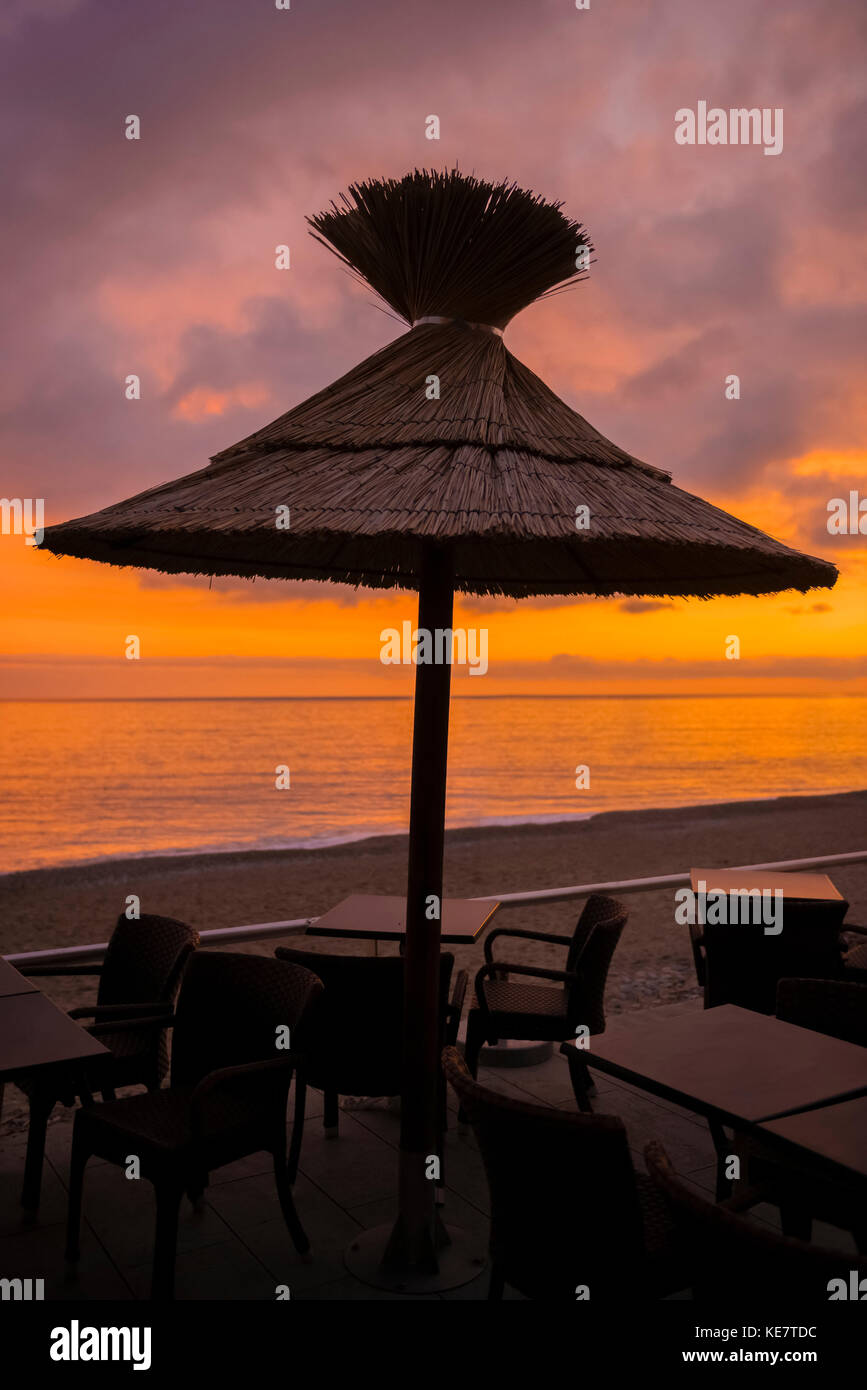 Un parasol en chaume sur une table avec des chaises sur la plage au coucher du soleil, avec vue sur la mer Méditerranée, Menton, Côte d'Azur, France Banque D'Images