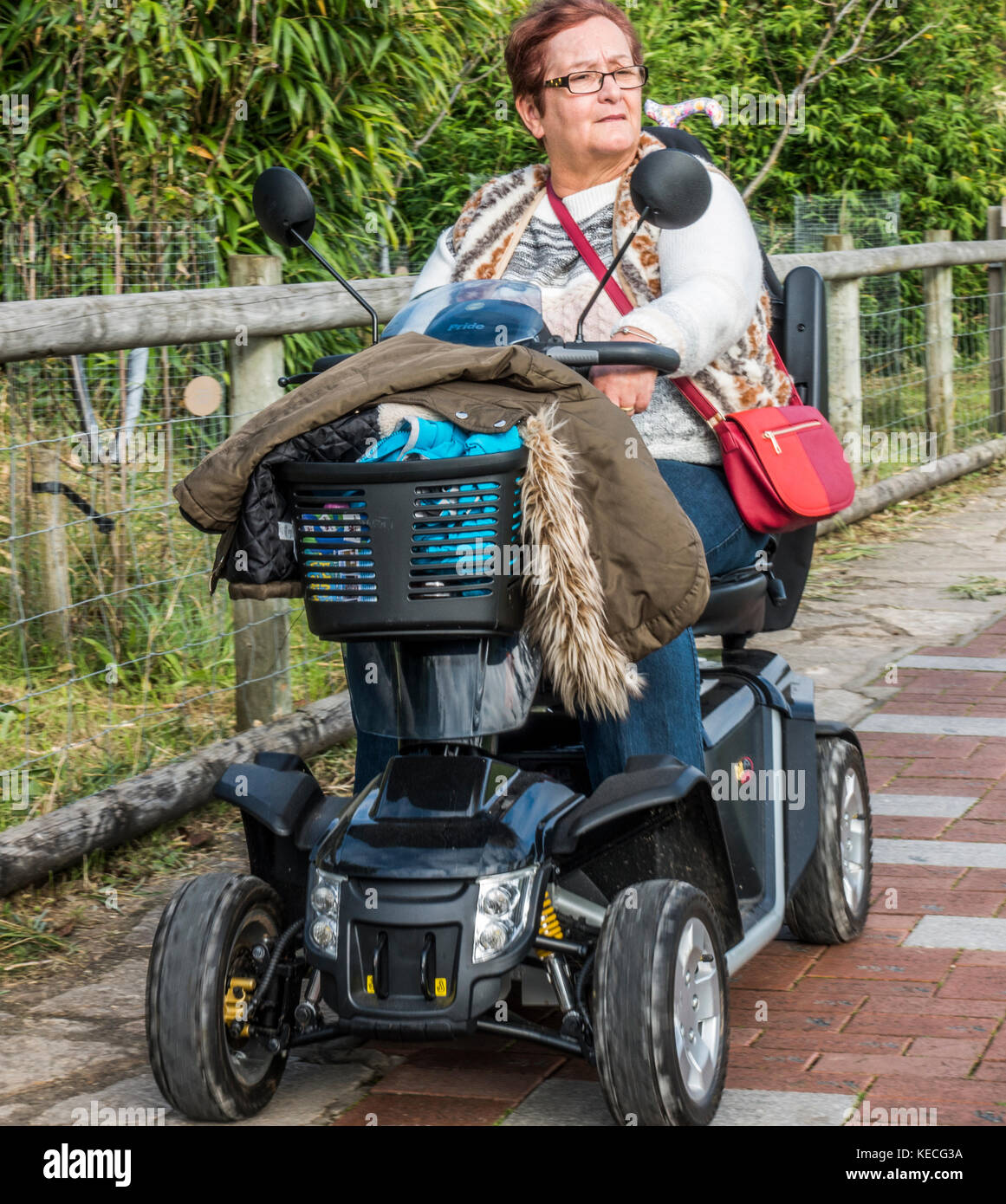 Une femme âgée d'une batterie de scooter de mobilité, avec des vêtements dans le panier avant et un sac rouge autour de son épaule. Dorset, Angleterre, Royaume-Uni. Banque D'Images