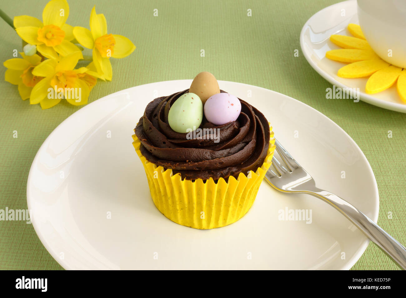 Libre d'un glaçage au chocolat cupcake de Pâques et les oeufs de pâques sur une plaque blanche avec des fleurs de narcisse et de café tasse sur fond vert. Banque D'Images