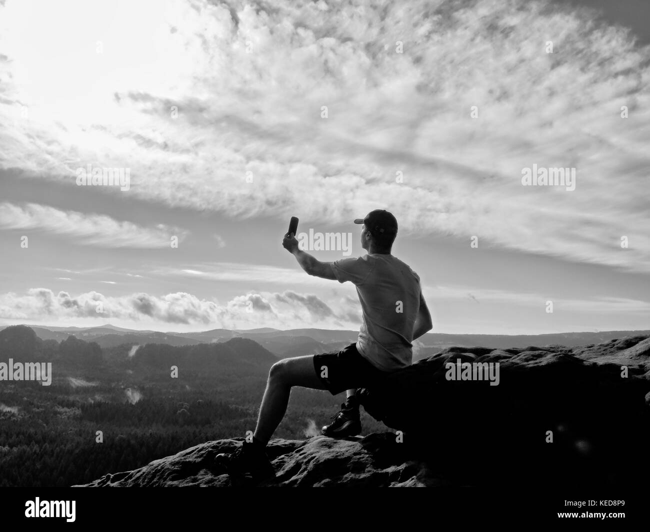 Touriste dans Grey T-shirt et casquette de baseball rouge prend des photos avec smart phone sur le pic de roche. paysage vallonné de rêve ci-dessous, le ressort misty lever du soleil Banque D'Images