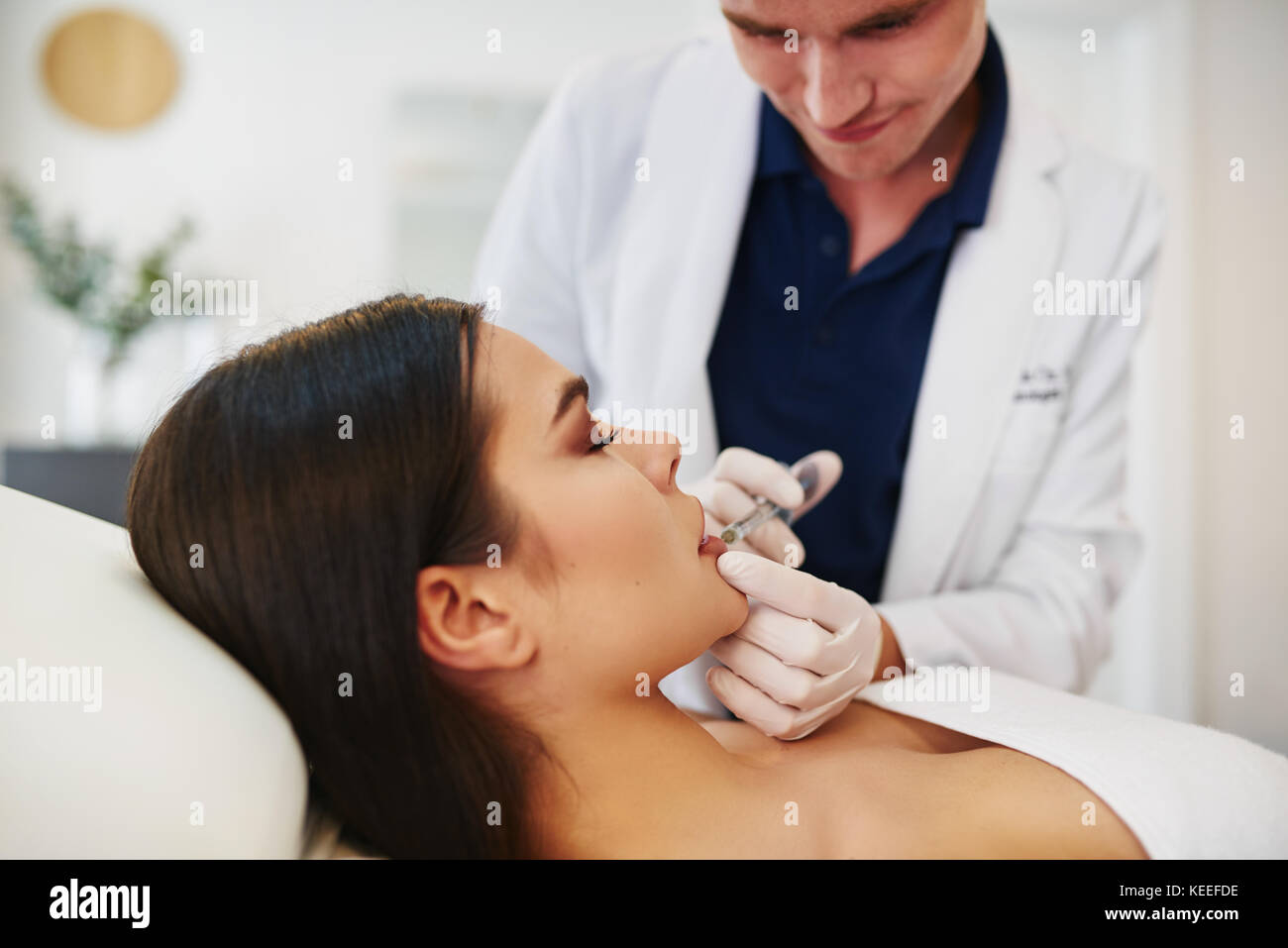 Jeune homme médecin d'effectuer des injections de botox pour les lèvres d'une femme brune allongé sur une table dans une clinique de beauté Banque D'Images