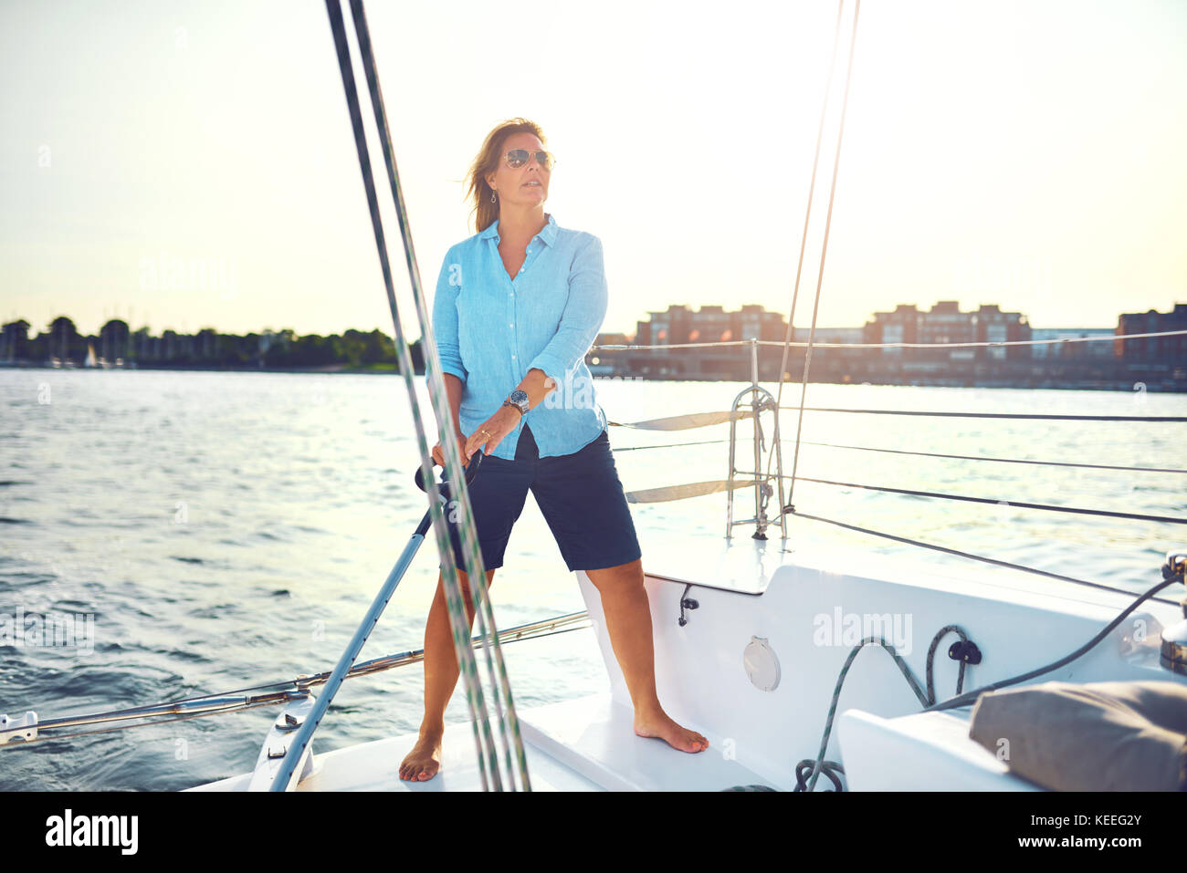 Mature Woman standing sur le pont d'un bateau avec la gouverne de direction tout en naviguant le long de la côte sur une journée ensoleillée Banque D'Images