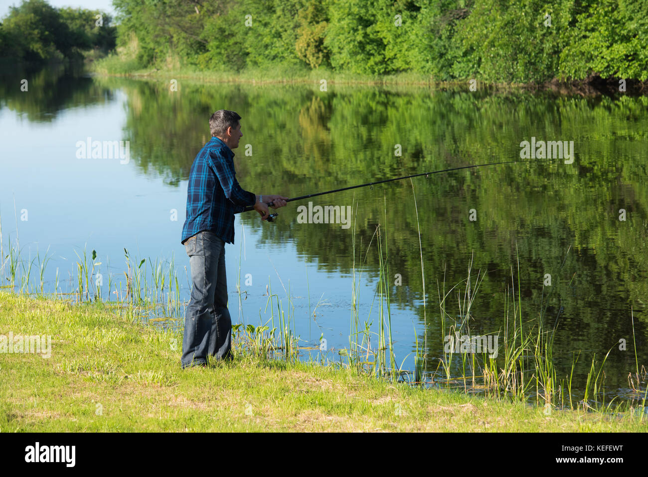 L'homme est à la retraite avec la pêche rod. Il est debout à la banque environ calme petite et étroite rivière. banque est couverte par l'herbe verte court. le ciel bleu Banque D'Images