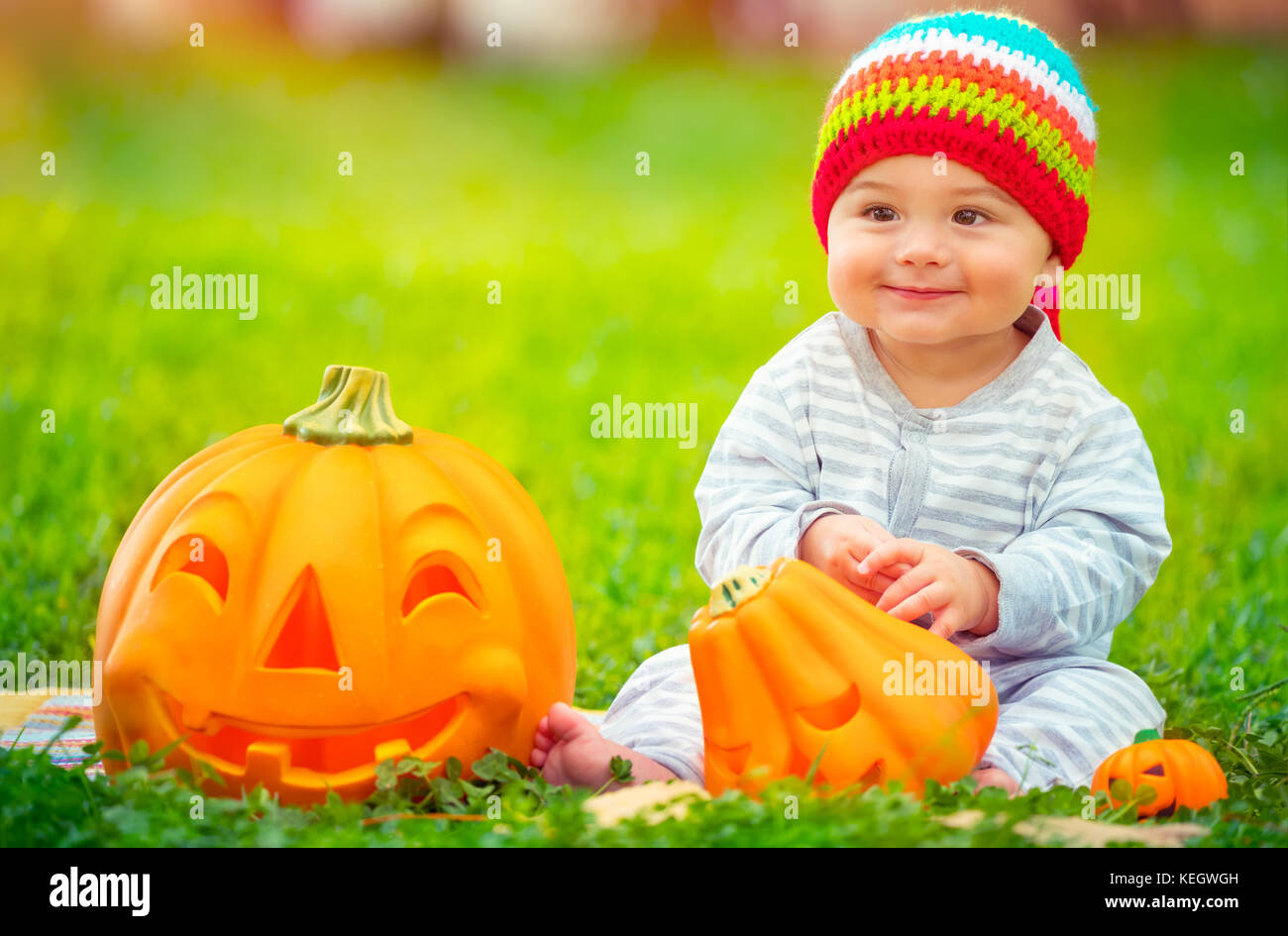 Cute little baby boy wearing colorful funny hat, assis dans la cour sur l'herbe verte prairie près de deux citrouilles décoratives, jeux en plein air avec traditi Banque D'Images