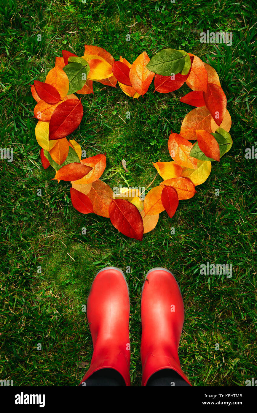 Portrait d'une femme en bottes en caoutchouc rouge à la forme de cœur à créé par feuilles mortes allongé sur l'herbe verte. Banque D'Images