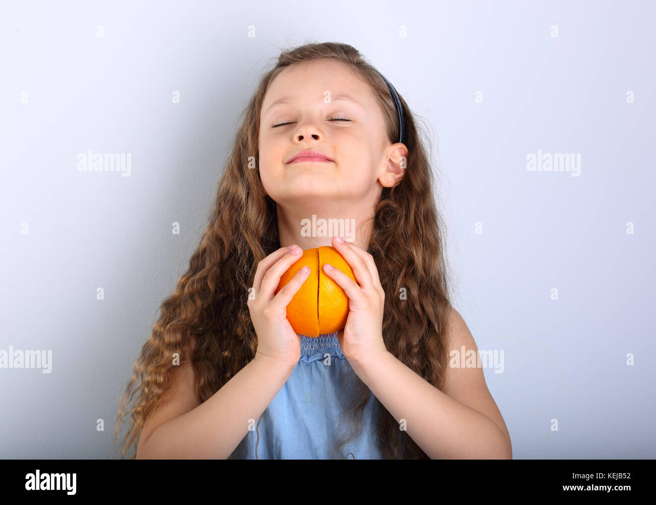 Cute smiling happy kid fille avec style les cheveux bouclés tenant ses mains les tranches de fruits orange agrumes avec sous les yeux ouvert bénéficiant sur fond bleu Banque D'Images